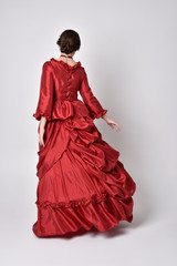 full length portrait of a brunette girl wearing a red silk victorian gown. Standing pose, with back...