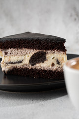 Creamy cheesecake with chocolate cookies and cream biscuits, chocolate cookies and cream biscuits cheese cake. Piece of cake on black plate, white cup on white marble background. Vertical photo.