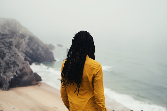 African male wearing long dreadlocks standing on ocean shore, white waves and cliffs in fog on background