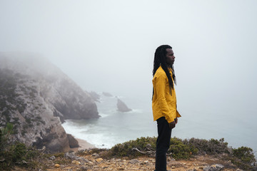 African male wearing long dreadlocks standing on ocean shore, white waves and cliffs in fog on...