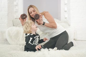 Mother and son child having fun with tasty sweet donuts indoors. Boy in halloween skeleton costume playing with lovely mom.