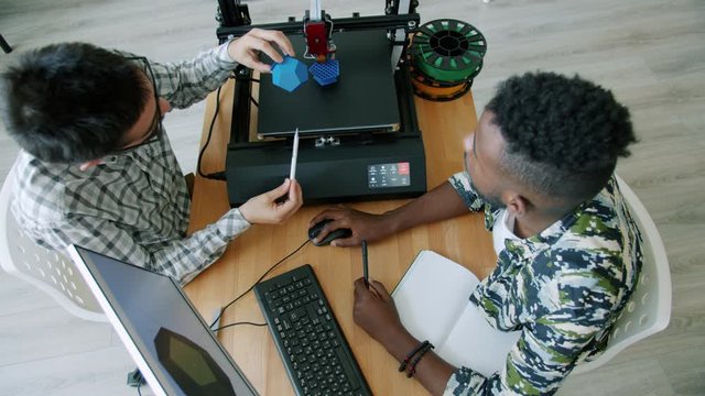 High angle view of smart guys making 3d model with three dimensional printer using computer in office talking discussing design. People and communication concept.