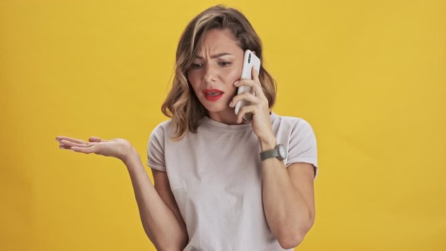 Displeased young woman with red lips expressing outrage while talking on mobile phone over yellow background isolated                                                                      