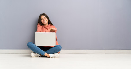 Young student girl with a laptop on the floor extending hands to the side for inviting to come