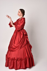 full length portrait of a brunette girl wearing a red silk victorian gown. Standing in side profile on a white studio background.