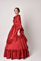 full length portrait of a brunette girl wearing a red silk victorian gown. Standing in side profile on a white studio background.