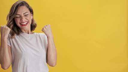 Smiling young woman with red lips showing a winner gesture over yellow background isolated                                                                          
