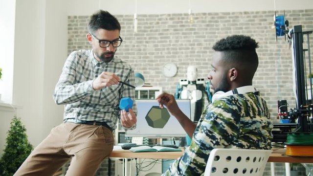 Young men Caucasian and African American are discussing 3d printed plastic element in office in workplace holding shape talking. Modern technology and business concept.