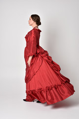 full length portrait of a brunette girl wearing a red silk victorian gown. Standing in side profile on a white studio background.