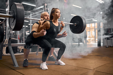 Naklejka na ściany i meble Pretty charming sports woman doing squats using heavy barbell, professional trainer standing behind, backing up, white smoke in the air, practicing in modern fitness center