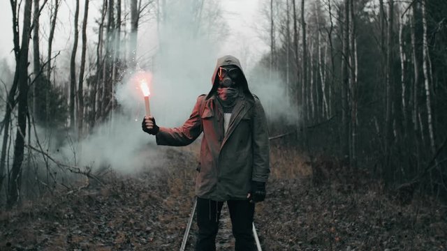 Man in gas mask standing on railway in empty dead forest holding red signal fire in hand. Armageddon concept, stalker after nuclear or chemical disaster. Post apocalyptic world. Slow motion