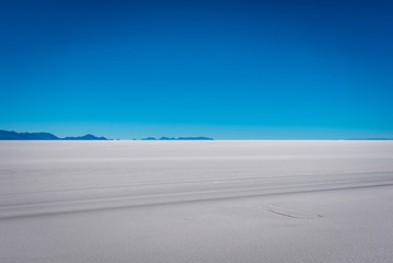 4×4 tracks cut across the Uyuni salt flats with mountains lining the horizon in the distance on a clear blue sky day.
