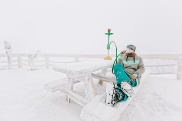 Snowboarder man relaxing and smoke a hookah or shisha, enjoying mountain view, winter frozen day,...
