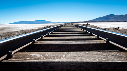 Wooden train tracks cross the Bolivian desert travelling towards the blue sky and distant mountains on the horizon.