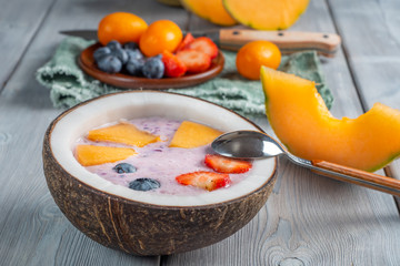 Close up view of blueberry smoothie in a coconut bowl with strawberries and melon and kumquat on a white wooden background with teaspoon and knife.. Soft focus.