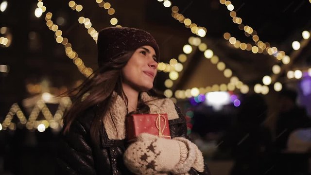 Young Cute Smiling Girl Wearing Winter Hat Holding Present Box, Looking Happy and Excited , Walking in the Falling Snow at Christmas Fair Lights Background. Holidays Concept.