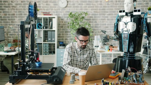 Young male scientist is working with robot, laptop and 3d printer in workplace developing cool gadgets at work. Innovation, technology and business concept.