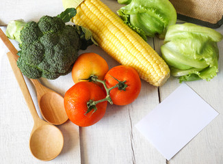 group of fresh and healthy vegetables, tomatoes, sweet corn, bloccoli with wooden spoons on white wooden table with white note paper,
