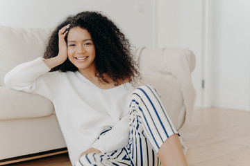 Lovely dark skinned woman with crisp hair, sits on floor near sofa, wears white sweater and striped...