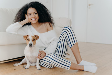 Horizontal shot of beautiful curly woman leans at sofa, sits on floor near pedigree dog, wears sweater, striped pants and socks. African American lady poses with pet. People and animals concept