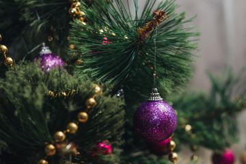 A small decorative Christmas ball covered with lilac spangles on a branch of a New Year's green tree decorated with gold beads, selective focus
