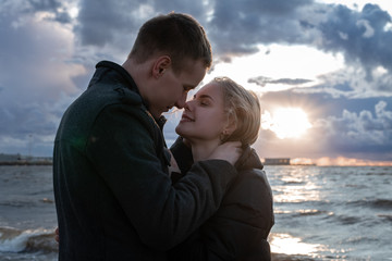 Young beautiful couple smiling happy face to face on the beach in cold autumn. Stunning sensual outdoor portrait of stylish and fashion couple posing on sunset. Happy to be in love.