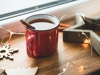 Black tea with cinnamon in a red mug among winter decor and lights. Cozy winter time still life.