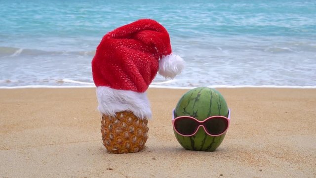 Picture of pineapple wearing Santa Claus hat and green cheerful watermelon in sunglasses on the tropical beach. New Year and Christmas concept.