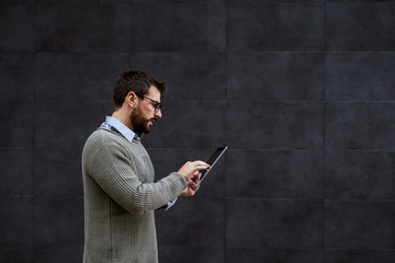 Side view of handsome bearded stylish caucasian man in gray sweater and with eyeglasses using tablet while passing by gray wall.