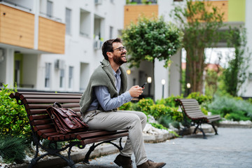 Fototapeta premium Side view of laughing caucasian stylish businessman sitting on bench after work and using smart phone. Next to him is handbag.