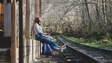 Young backpacker woman in denim overall is sitting on country train station alone with bottle of...