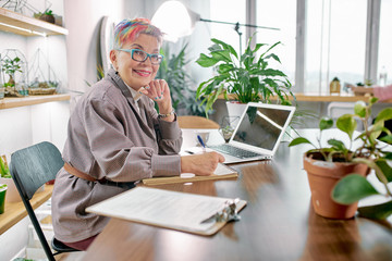 Charming modern short-haired woman dressed in neat blazer sit affably smiling and look at camera. documents, laptop and plants on table.