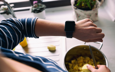 The woman is cooking the food in her kitchen and looking on her smartwatch on her hand