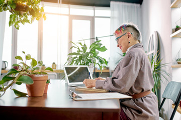 Side view on attractive caucasian female in glasses and short colorful hair sit on office table using laptop while working, plants and documents on table. Sunny day, light office