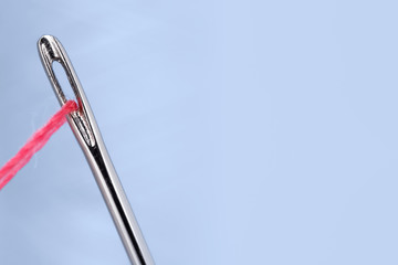 Steel sewing needle with a contrasting pink thread threaded through the eye. In the background is a spool of thread of blue color. Right place for text.