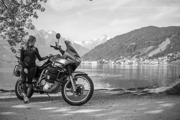 Obraz na płótnie Canvas Pretty women is standing with traveler motorcycle. Vacation and hobby concept, jorney on two wheels. Sunny summer day in the Alpine mountains. Zell am see lake on background Austria. Black and white