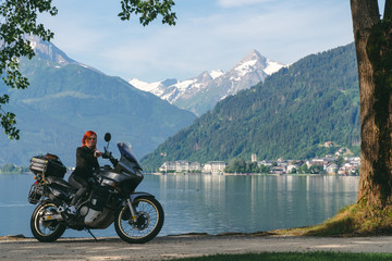 Fototapeta na wymiar Pretty women is sitting with traveler motorcycle. Vacation and hobby concept, jorney on two wheels. Sunny summer day in the Alpine mountains. Zell am see lake on background Austria. Copy space