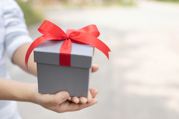 female hand holding gift box gray wrapped pack with beautiful ribbon red outdoor background. for surprise special event with copy space. Christmas,birthday or new year concept. High resolution product
