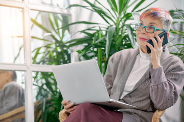 Strict middle-aged business lady boss working in office talking on mobile phone, with laptop