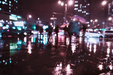 Blurred modern city at rayny night, defocused urban bokeh city lights, silhouettes of the walking people under umbrellas. Abstract background