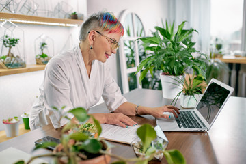 Modern florist have short colorful hairstyle, look at screen of laptop and typing. Different plants around, in room and on table