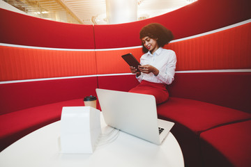 A young charming cheerful African-American business lady is sitting in a cozy office lounge zone on a curved red sofa and using her tablet pc, a laptop and a dock station on the table in front of her