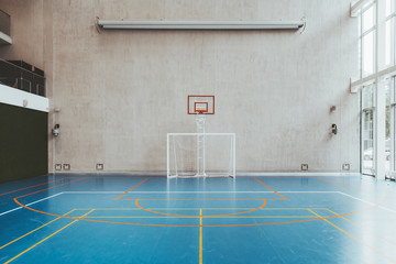 Obraz na płótnie Canvas Front view of the court in the gymnasium hall; an indoor modern office stadium with a basketball basket and hoop, football goal, blue floor, a concrete wall with an undeployed projection screen above