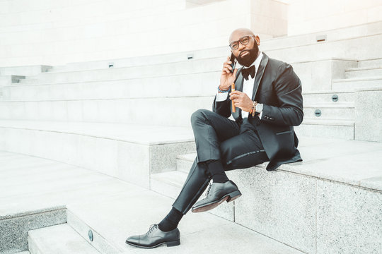 An Elegant Bald Afro Man In A Black Formal Suit And Spectacles, With A Beard And Holding A Cigar In His Hand, Is Sitting Outdoors On The Marble Seat Of An Amphitheater And Having A Phone Conversation