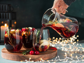 Female hand pours winter sangria in glasses with fruit slice, cranberry and rosemary. Dark christmas holiday background with candle, decoration lighting chain.
