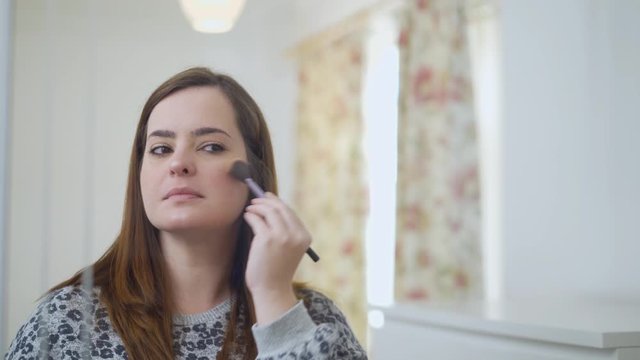 Woman applying Illuminator Powder with a feather-light in front of the mirror. Caucasian female using beauty cosmetics to improve herself ready to work in bedroom at home.