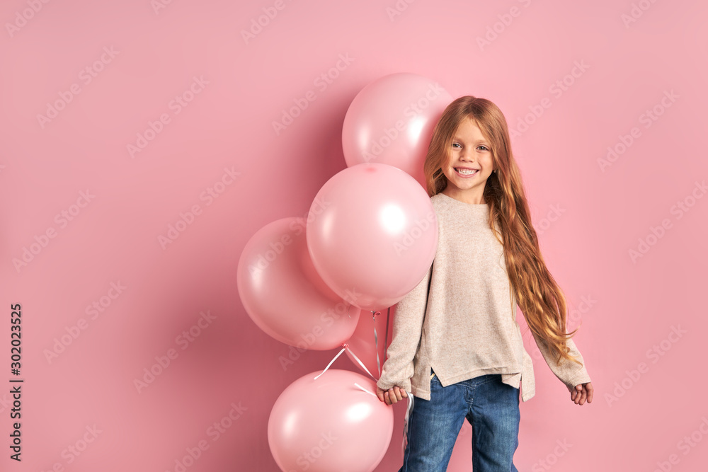 Wall mural Smiling caucasian girl with long hair isolated over pink background, holding bunch of pink air balloons. The concept of children holiday birthday