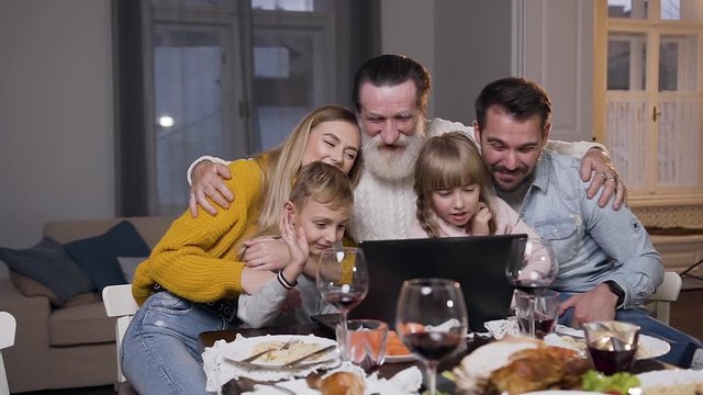 Attractive different generation family having video chat on computer during festive dinner and greeting their interlocutor with waving hands