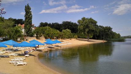Fototapeta na wymiar Cozy summer lake beach with blue umbrellas on the green coast