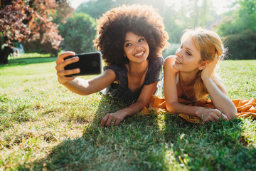 Couple of young women take a selfie with the smart phone lying on the grass of a meadow on a summer...